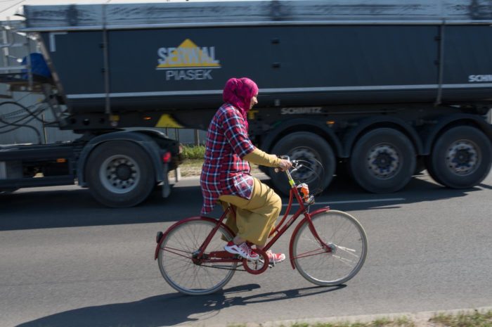 Na zdjęciu uchodźczyni na rowerze w chuście, poruszająca się rowerem po jezdni, wymija ciężarówkę